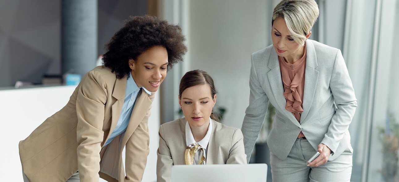 Three professional women collaborating around a laptop in a modern office setting, discussing digital product design and IT management for Carolina Hyperbaric & Wellness projects.