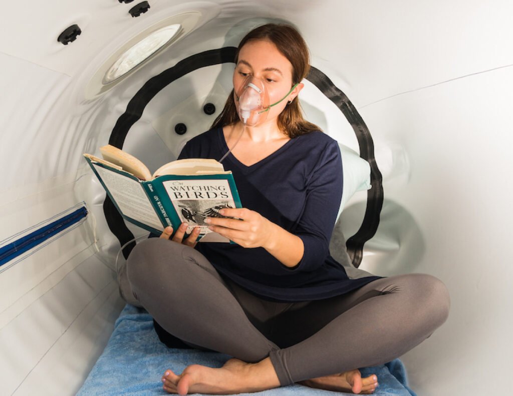 Woman reading a book inside a hyperbaric chamber, wearing a mask for oxygen therapy, emphasizing relaxation and wellness during the session.